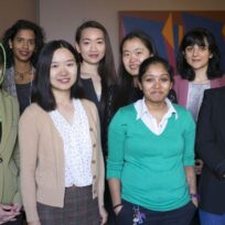A group of eight women stand together indoors, facing the camera and smiling. A colorful abstract painting is visible in the background.