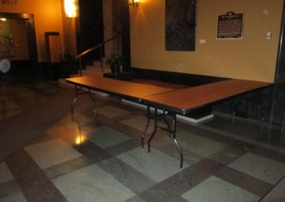 A large rectangular table with folding legs placed against a wall in the Rackham Building lobby with checkered flooring.