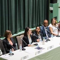 Seven panelists sit at a long table with microphones, water bottles, and nameplates, engaged in a discussion in front of a green curtain.