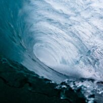 Inside view of a large ocean wave forming a tunnel with swirling water patterns. The light blue and white colors create a dynamic and powerful image.
