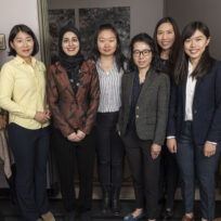 Eight women stand in a row indoors, dressed in business or smart casual attire, facing the camera and smiling.