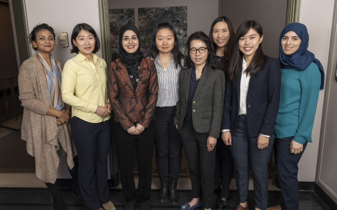 Eight women stand in a row indoors, dressed in business or smart casual attire, facing the camera and smiling.
