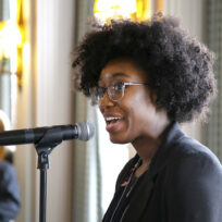 A person with curly hair and glasses speaks into a microphone at an indoor event, with people and tall windows visible in the background.