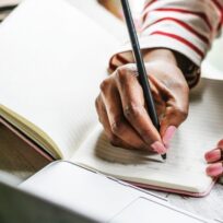 Person writing in a notebook with a pen. Nearby, a laptop and a pair of sunglasses are visible. The person is wearing a striped shirt.