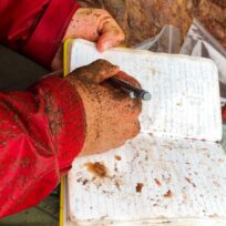 Person with muddy hands writes in a notebook outdoors, wearing a red jacket and sitting next to a stone surface.