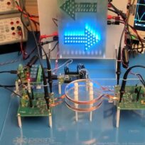 A laboratory setup with electronic components, oscilloscopes displaying waveforms, and illuminated blue arrows pointing right. Various cables and circuit boards are connected on a blue table.