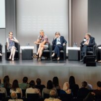 Panel of six people seated on stage having a discussion, with an audience watching.
