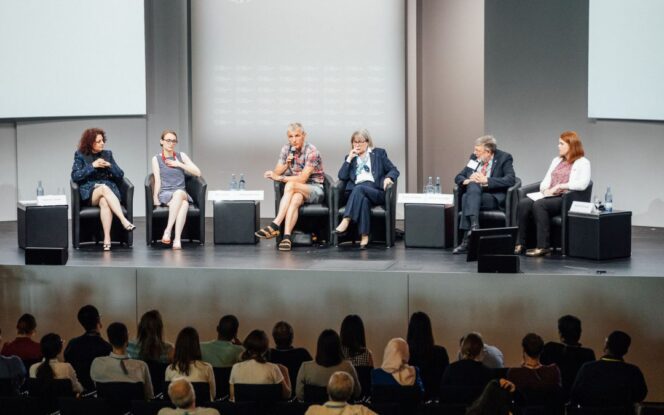 Panel of six people seated on stage having a discussion, with an audience watching.