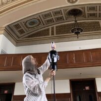 A person adjusts a mannequin head with microphones on a stand in a room with ornate ceilings and wood paneling.