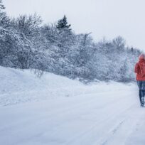 Person in a red jacket walking on a snow-covered road, surrounded by snow-laden trees under a cloudy sky.