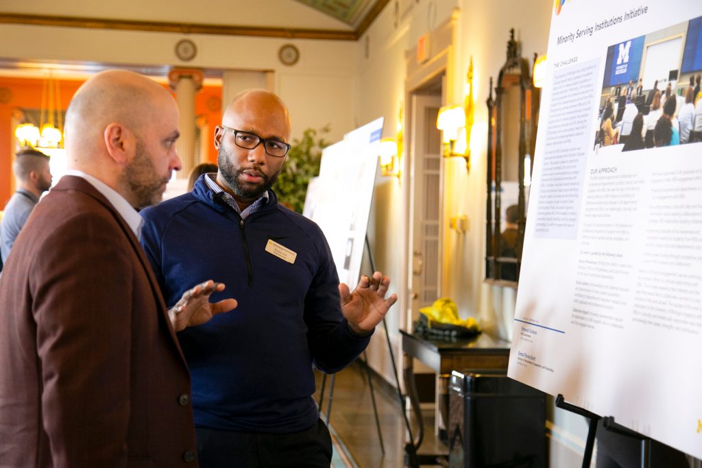 Two men in conversations in front of a poster labeled "Minority Serving Institutions Initiative."