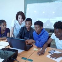 A group of five people gather around a laptop at a table, with papers and a whiteboard in the background.