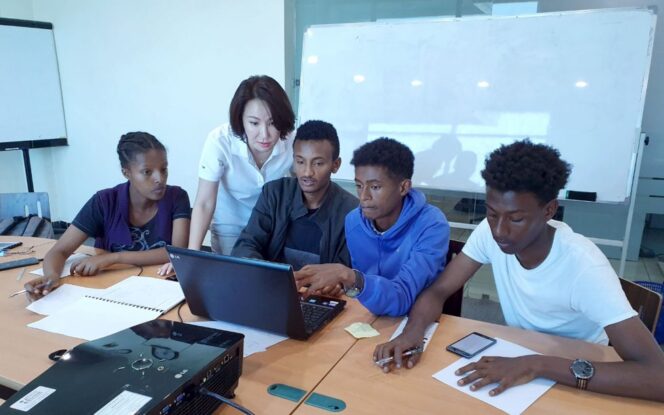 A group of five people gather around a laptop at a table, with papers and a whiteboard in the background.