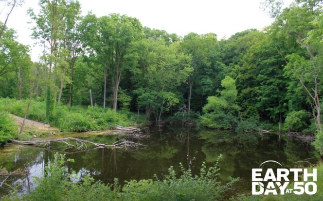 A tree-lined pond with text reading "Earth Day at 50" in the lower right corner.