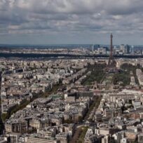 Aerial view of Paris featuring the Eiffel Tower at the center with buildings and streets surrounding it. Cloudy sky and distant cityscape visible in the background.
