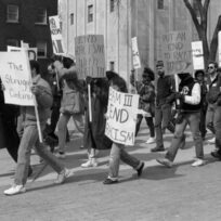People marching with signs protesting racism, including messages like "End Racism" and "The Struggle Continues," on a street lined with buildings.