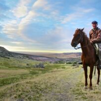 A man wearing a jacket and cap sits on a brown horse in a grassy, open landscape with hills under a partly cloudy sky.