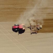 A red tractor pulls farming equipment across a dry, brown field, creating dust as it tills the soil.