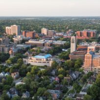 Aerial view of a city campus with academic buildings, residential houses, and tree-lined streets under a clear sky.