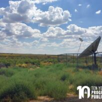 A satellite dish stands in a grassy field near a chain-link fence under a partly cloudy sky; text reads "Ten Years Program in Public Scholarship.