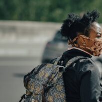 A person wearing a patterned face mask and backpack stands outdoors near a road, with blurred cars in the background.