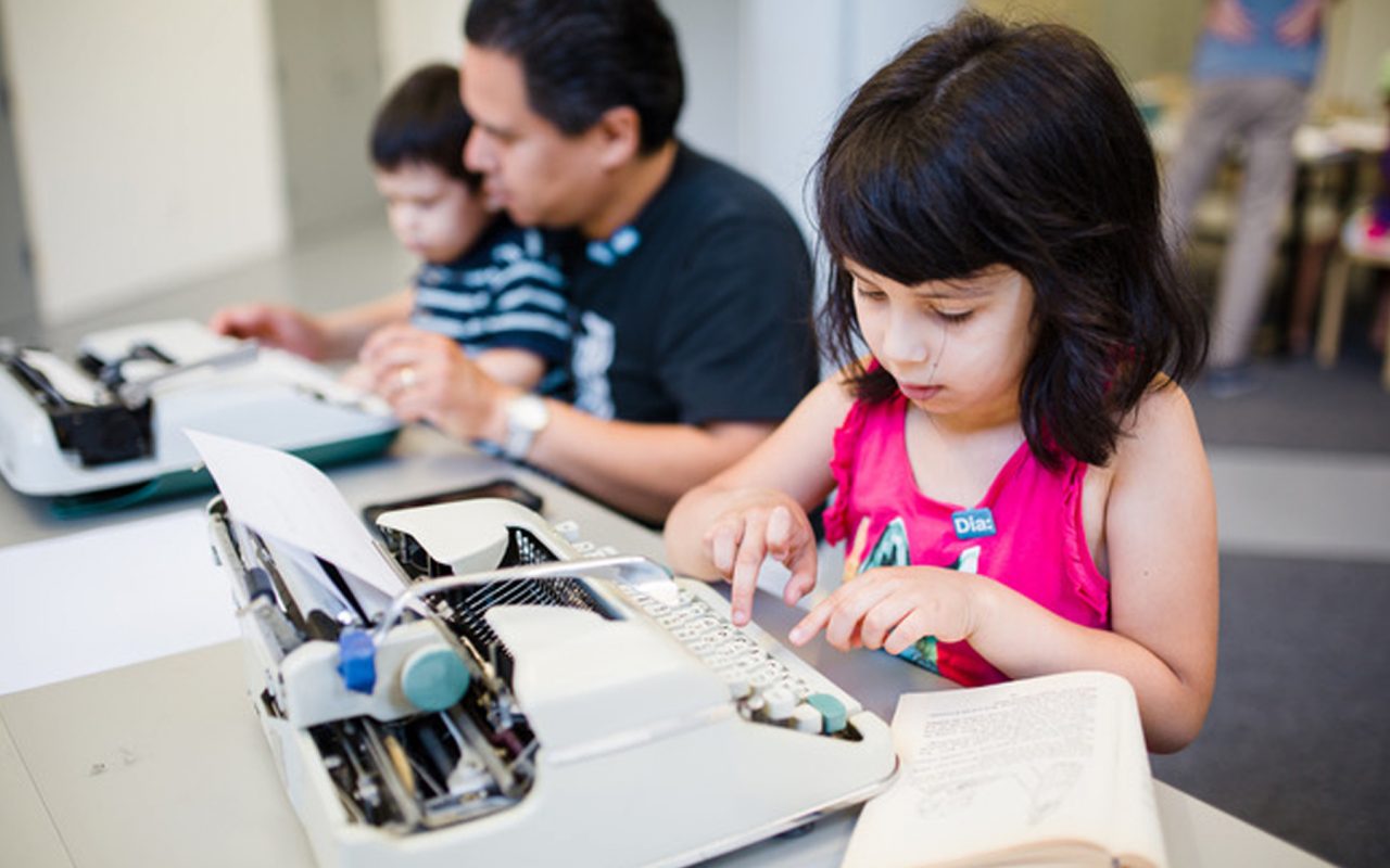 A young girl using a typewriter, with a man and a young boy using another typewriter in the background.