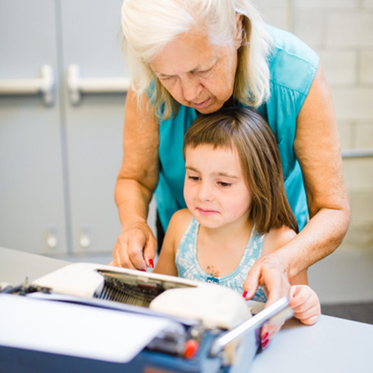 An older woman helping a young girl type on a vintage typewriter.