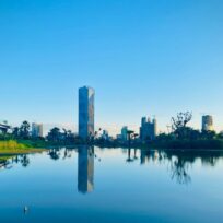 Cityscape with modern buildings reflected in a calm body of water, surrounded by greenery under a clear blue sky.