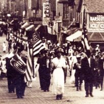 A black and white photo shows a parade on a city street with people carrying American flags and musical instruments, while crowds line the sidewalks.