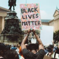 A person in a crowd holds up a colorful sign reading "Black Lives Matter" during a protest in front of a large, classical building.