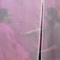 Two people wearing glasses and face masks sit facing each other, separated by a sheer pink curtain, with one person extending their hand forward.