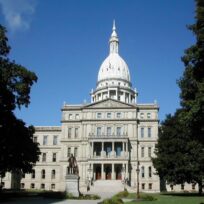 The Michigan State Capitol building in Lansing, featuring a white dome, classical architecture, and a statue in front, set against a clear blue sky and framed by trees.