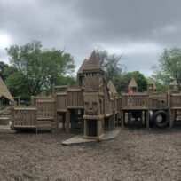 A large wooden playground structure with towers, bridges, slides, and ladders, set outdoors on a cloudy day with trees in the background.