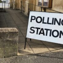 A large white sign reading "POLLING STATION" is displayed outside a building near a paved walkway and stone wall.