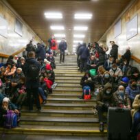 A large group of people sit and stand on the steps of an indoor stairwell, many with bags and some wearing masks, appearing to wait or take shelter.