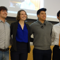 Four people stand together indoors, smiling. One person holds a microphone. A screen in the background shows a video call.