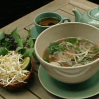A bowl of Vietnamese pho with beef, accompanied by fresh herbs, bean sprouts, lime, chili, and a pot of tea on a wooden table.