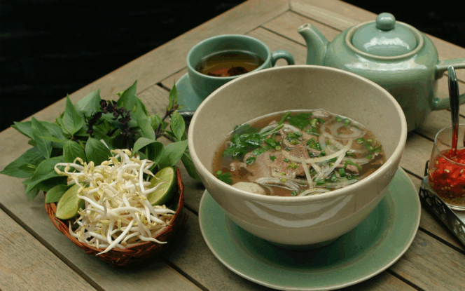 A bowl of Vietnamese pho with beef, accompanied by fresh herbs, bean sprouts, lime, chili, and a pot of tea on a wooden table.