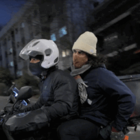 Two people ride a motorcycle at night; the driver wears a helmet and the passenger wears a white beanie and scarf, holding onto the driver's waist.
