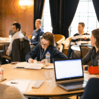 People sit around tables in a conference room, some listening and some taking notes. Laptops, notebooks, and drinks are on the tables.