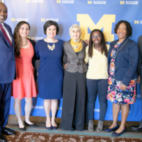 Seven people standing in front of a blue backdrop with the University of Michigan logo, wearing formal attire, and smiling.