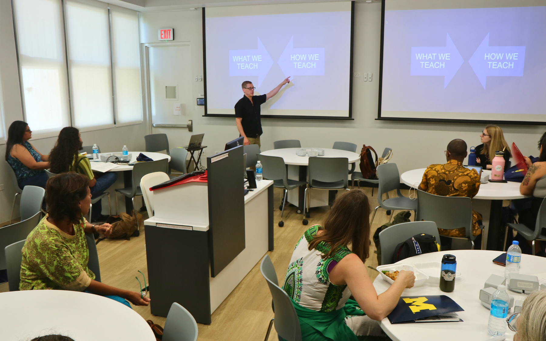 An individual stands at the front of a classroom, pointing at a power point presentation.