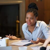 A woman speaks during a meeting with three others seated around a table. A sign reads "Community Action Network." Papers and cups are on the table.