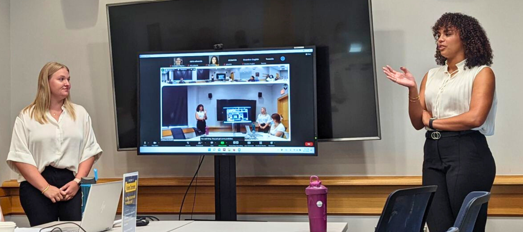 Sayler presents to a hybrid audience in-person and via Zoom. A large screen with her Zoom presentation is behind her. 