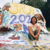 A person is sitting in front of a painted rock with colorful designs and the text "SRP 2022" and "#HereToStay" outdoors.
