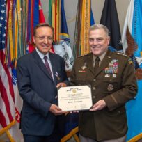 Two men stand smiling; one is in a military uniform holding a certificate with the other in a suit. Several flags are displayed in the background.