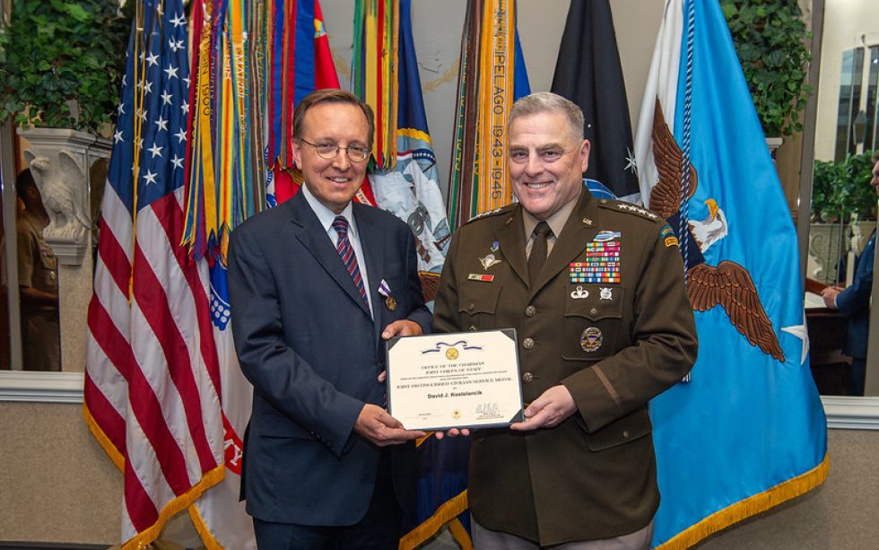 Two men stand smiling; one is in a military uniform holding a certificate with the other in a suit. Several flags are displayed in the background.