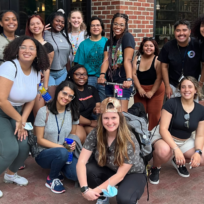 A group of people poses and smiles in front of a brick building. Some are kneeling, others standing, holding water bottles.