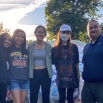 Five people standing outdoors smiling, with trees and a painted rock in the background. One person is wearing a face mask.