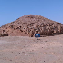 A small, weathered pyramid made of stone blocks under a clear blue sky, with a sign in front of it on a sandy terrain.